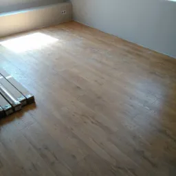 Ułożona podłoga z paneli drewnopodobnych w pokoju na poddaszu, z paczkami paneli w lewym dolnym rogu i oknem w tle.