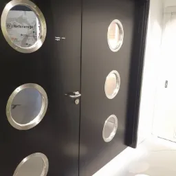 Czarne, dwuskrzydłowe drzwi wewnętrzne z okrągłymi okienkami w srebrnych ramkach, jedno z okienek zawiera napis 'Referencje' i mapę, widoczne na białej podłodze.