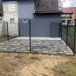 Wykończona powierzchnia z szarej kostki brukowej o wzorze rombów, otoczona metalową siatką ogrodzeniową na tle budynku z szarą elewacją i częściowo odkrytą cegłą.