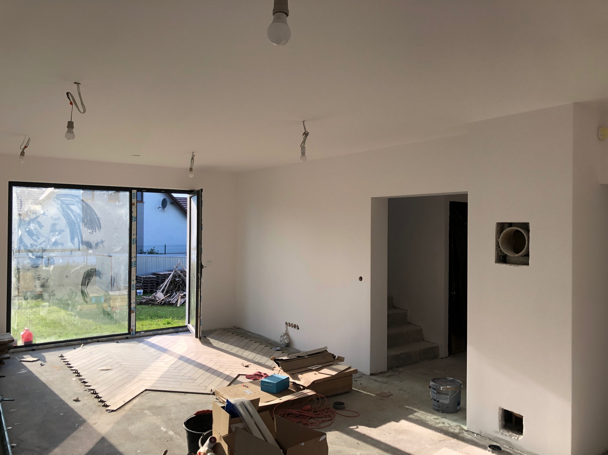 Wnętrze w trakcie remontu: świeżo pomalowane ściany na biało, częściowo ułożona podłoga w jodełkę, widoczne instalacje elektryczne, okno tarasowe z widokiem na ogród i materiały budowlane na podłodze.