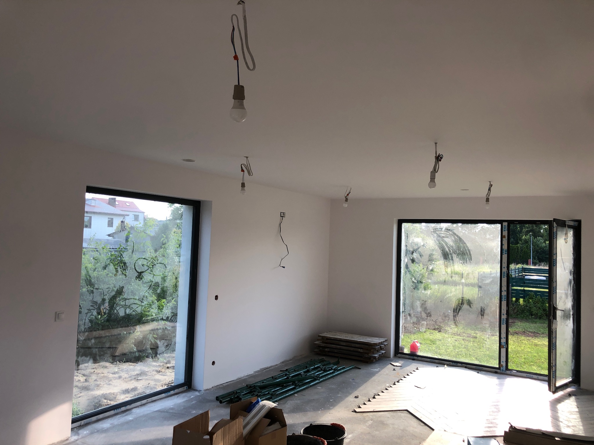 Wnętrze w trakcie prac wykończeniowych: surowe, białe ściany, tymczasowe oświetlenie, rozłożone rusztowanie, widoczne elementy podłogi i duże okna z widokiem na zieleń.