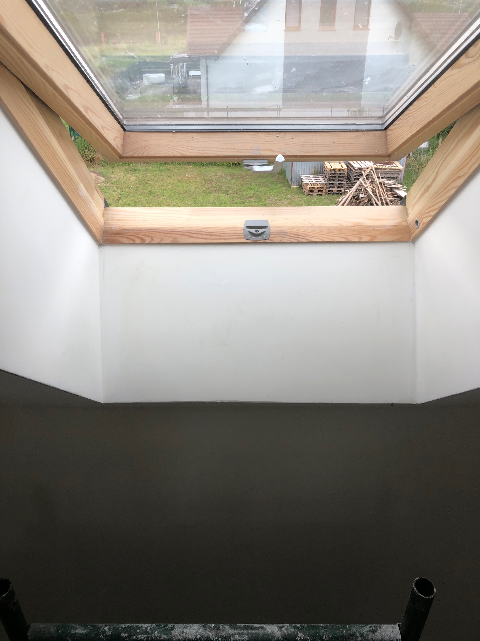 Wnętrze poddasza z oknem dachowym po szpachlowaniu ścian, widoczne palety i drewno na zewnątrz.