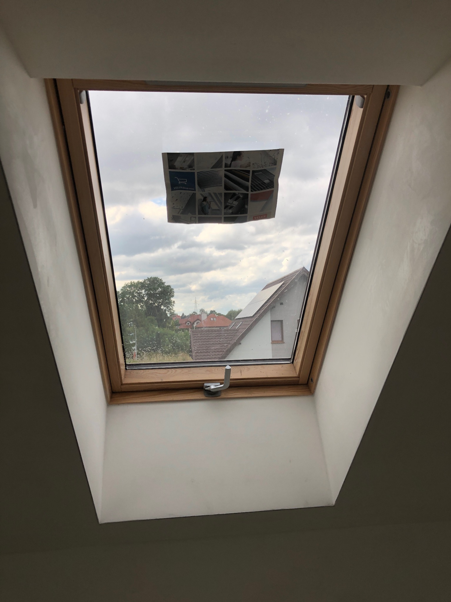 Świeżo wykończone poddasze z oknem dachowym, na szybie naklejona ulotka informacyjna, ściany pomalowane na biało, widok na sąsiednie domy.