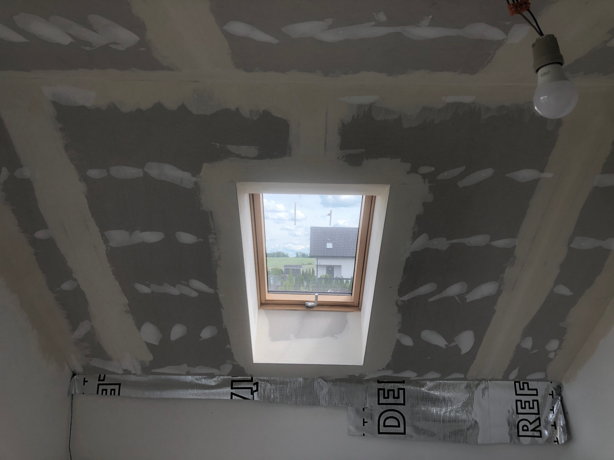 Poddasze w trakcie prac wykończeniowych: widoczne łączenia płyt kartonowo-gipsowych na skosach dachu, wstępnie pokryte masą szpachlową, okno dachowe z widokiem na dom i zieleń, folia paroizolacyjna...