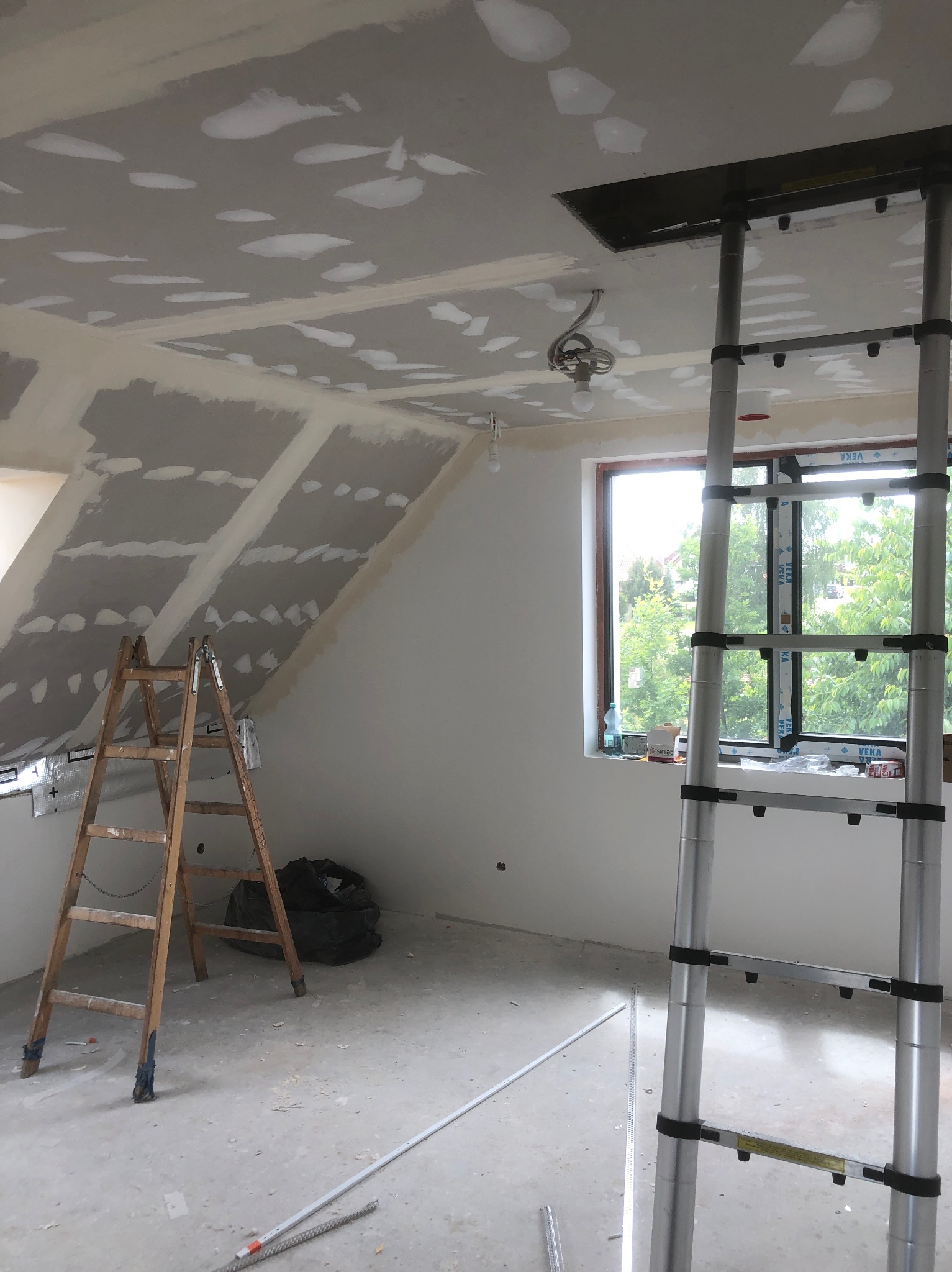 Wnętrze poddasza w Olsztynie w trakcie remontu: widoczne płyty karton gips na skośnym suficie z nałożoną szpachlą, dwie drabiny (drewniana i aluminiowa rozsuwana) oraz okno z widokiem na zieleń.