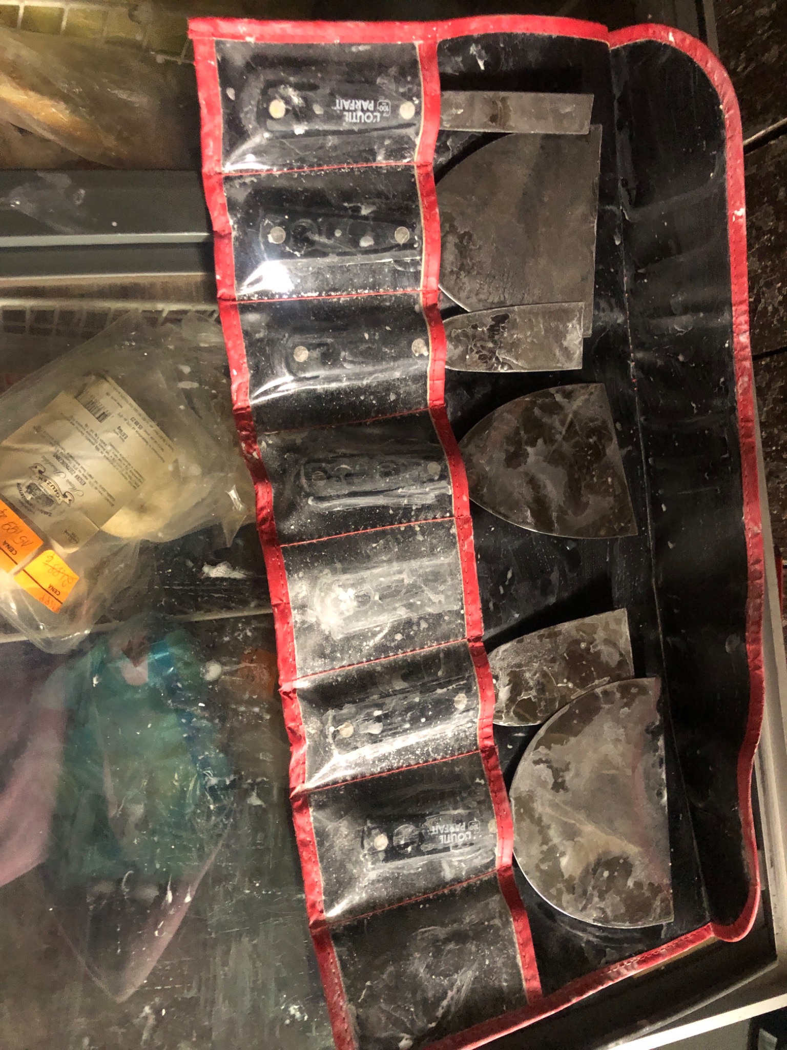 Zestaw używanych szpachelek w pokrowcu z czerwonymi brzegami, obok worek z tworzywa sztucznego z etykietami i resztkami materiałów budowlanych.