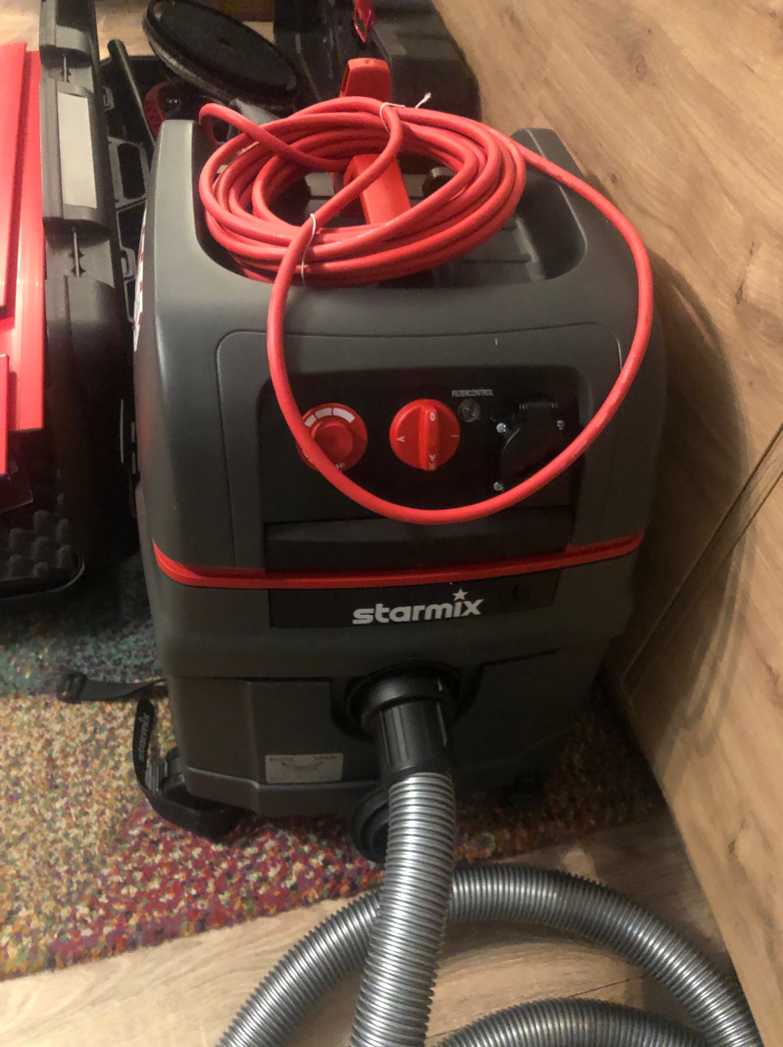 Profesjonalny odkurzacz przemysłowy Starmix z czerwonym kablem zwiniętym na górze, gotowy do użycia na tle drewnianej ściany i wzorzystego dywanu.