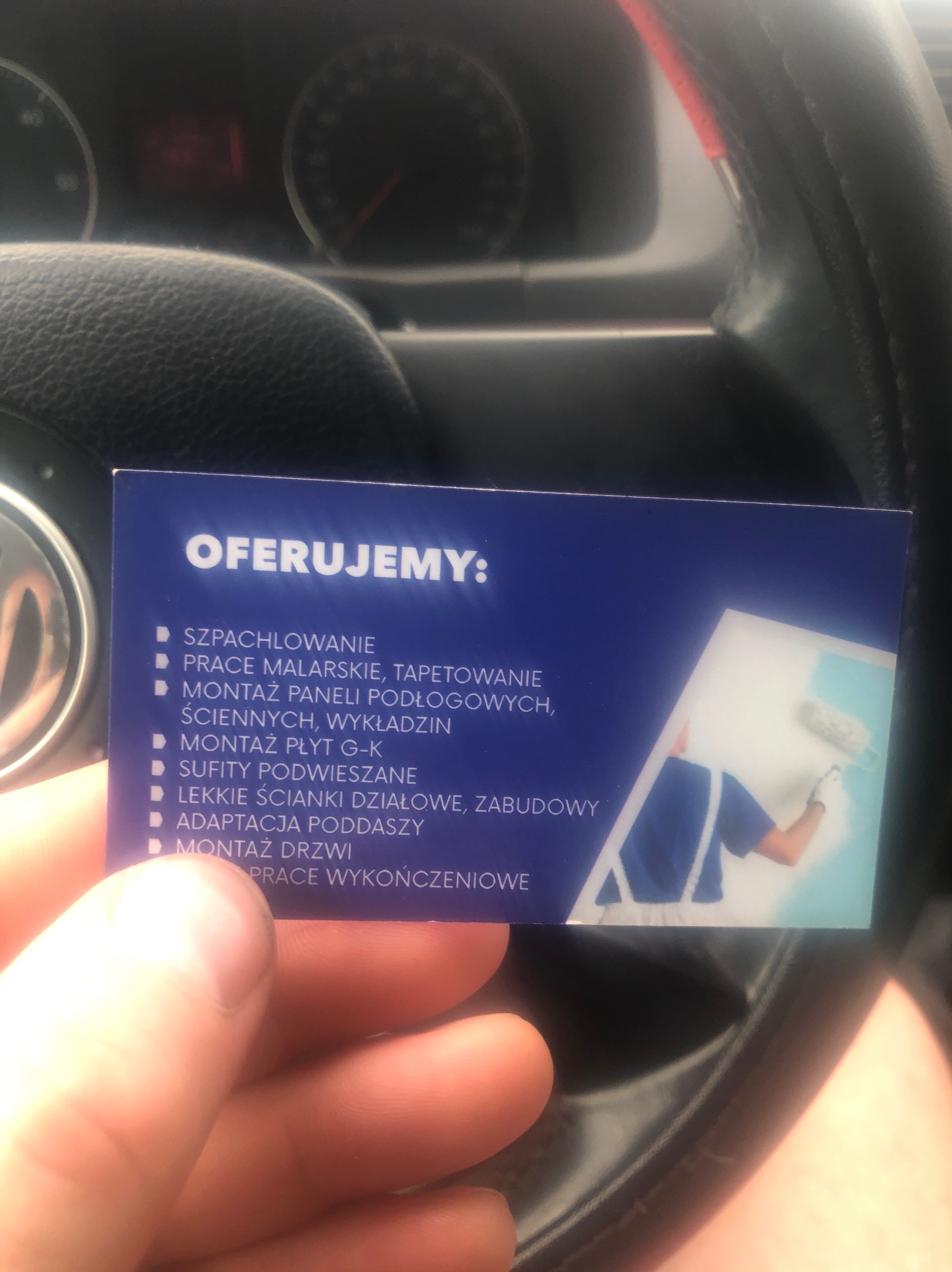 Wizytówka firmy remontowej trzymana w dłoni, prezentująca ofertę usług: szpachlowanie, prace malarskie, montaż płyt G-K, sufity podwieszane i inne prace wykończeniowe. W tle widoczna kierownica...