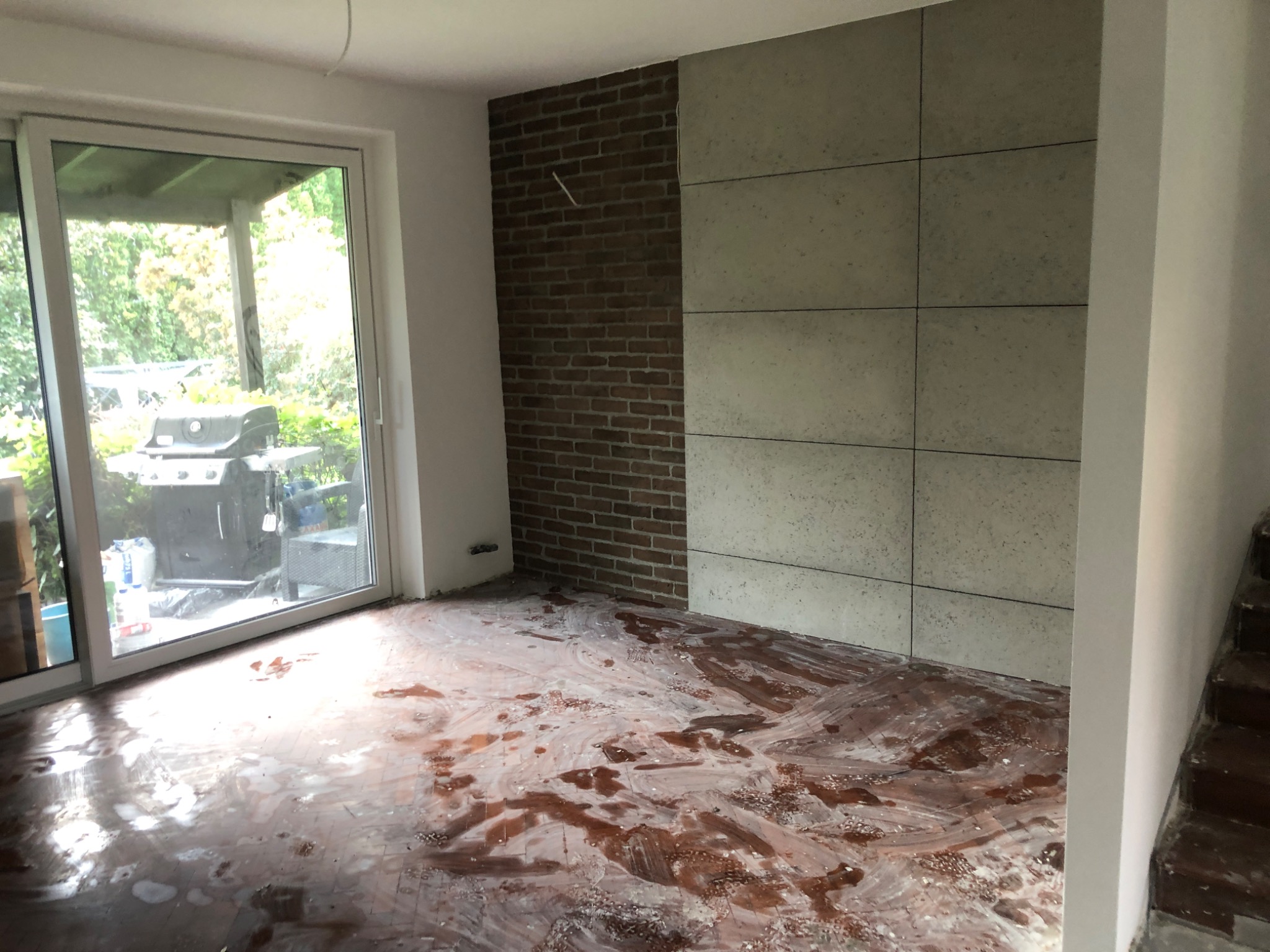 Pokój w trakcie przygotowania do prac wykończeniowych: podłoga z resztkami kleju lub farby, ściana z cegły i płyt betonowych, widoczne okno tarasowe.