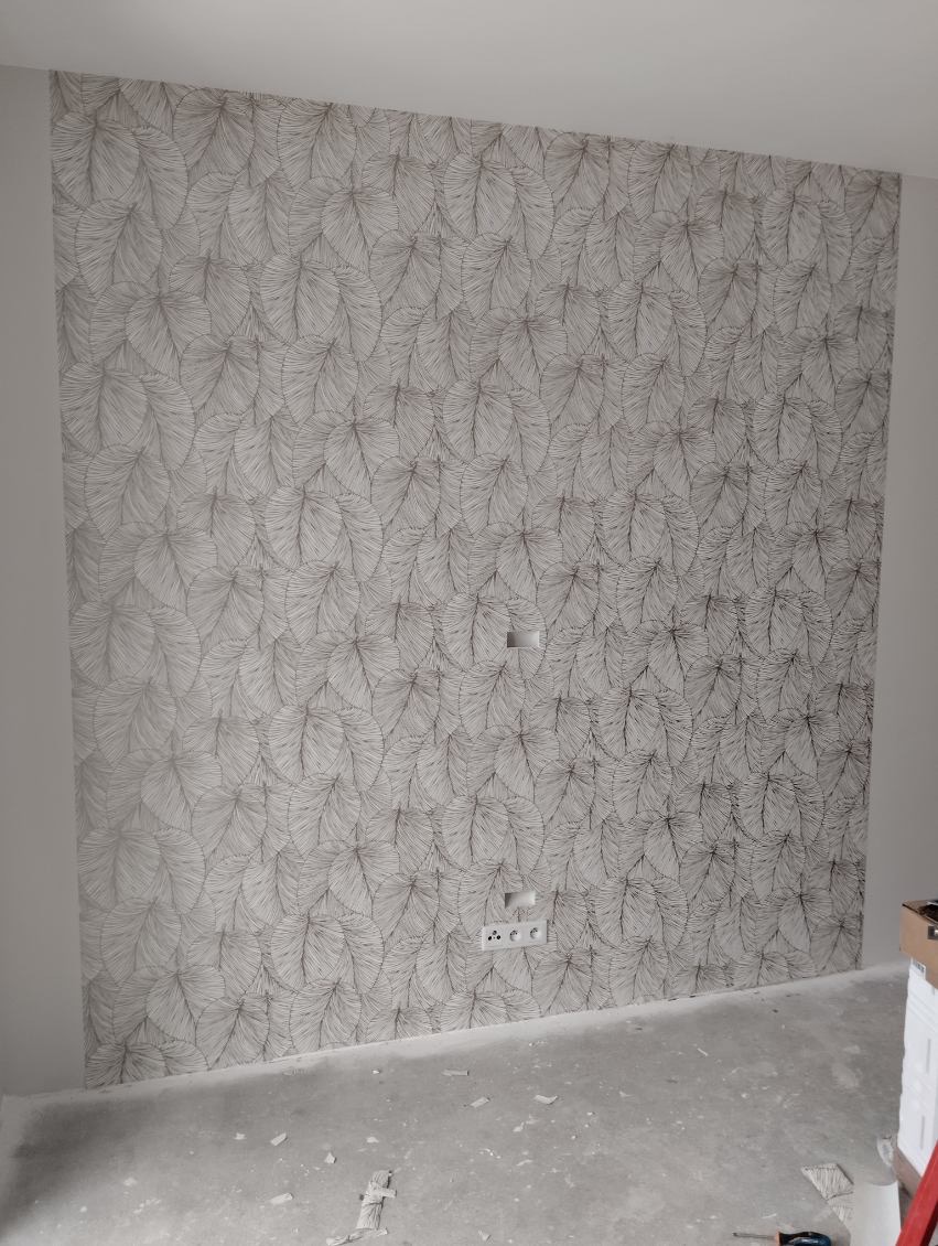 Ściana pokryta tapetą z wzorem liści, widoczne gniazdka elektryczne, surowy beton na podłodze.