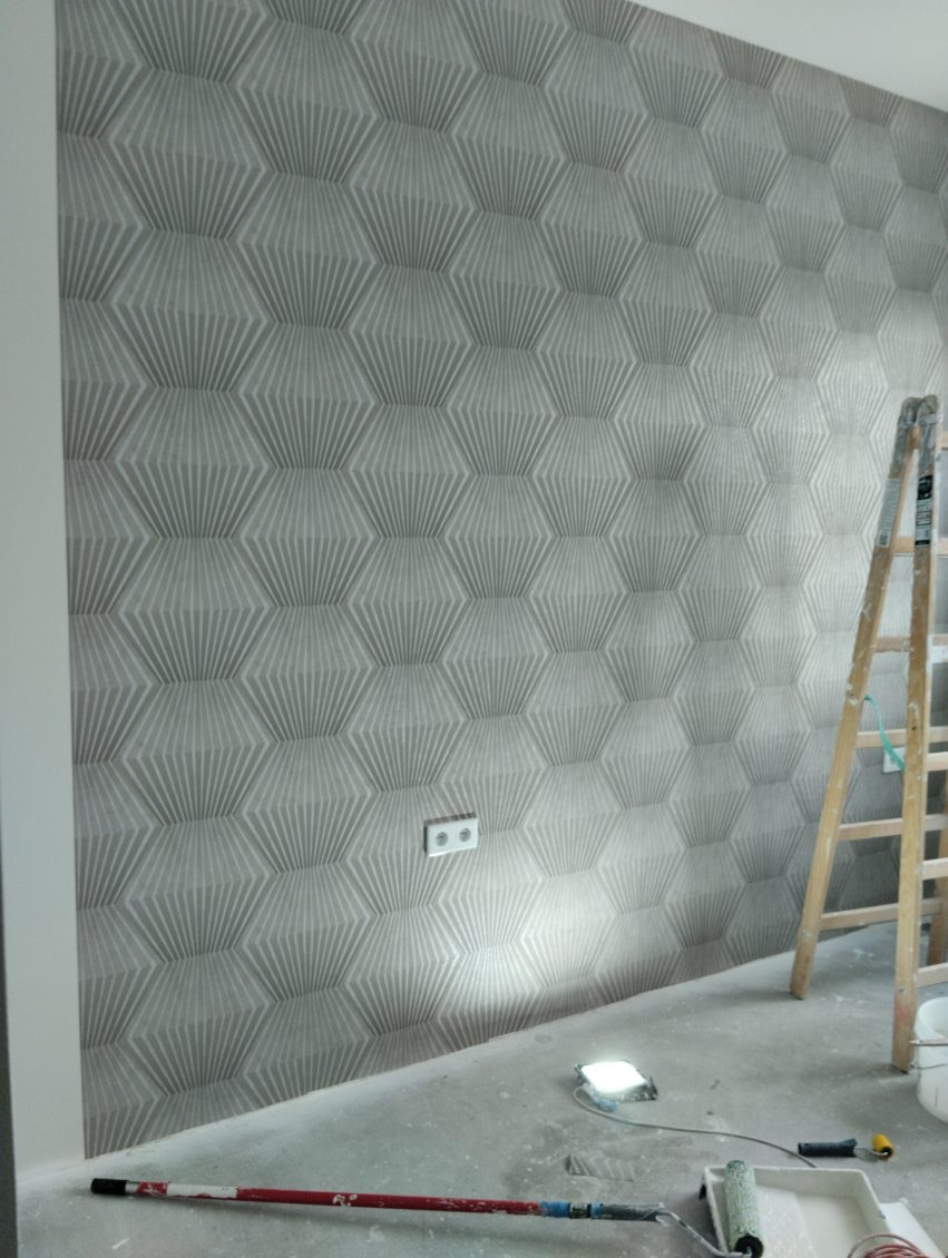 Ściana pokryta szarą, geometryczną tapetą 3D w kształcie heksagonów z widocznym gniazdkiem elektrycznym, drabina oparta o ścianę, wałek do malowania i kuweta na podłodze, oświetlenie robocze...