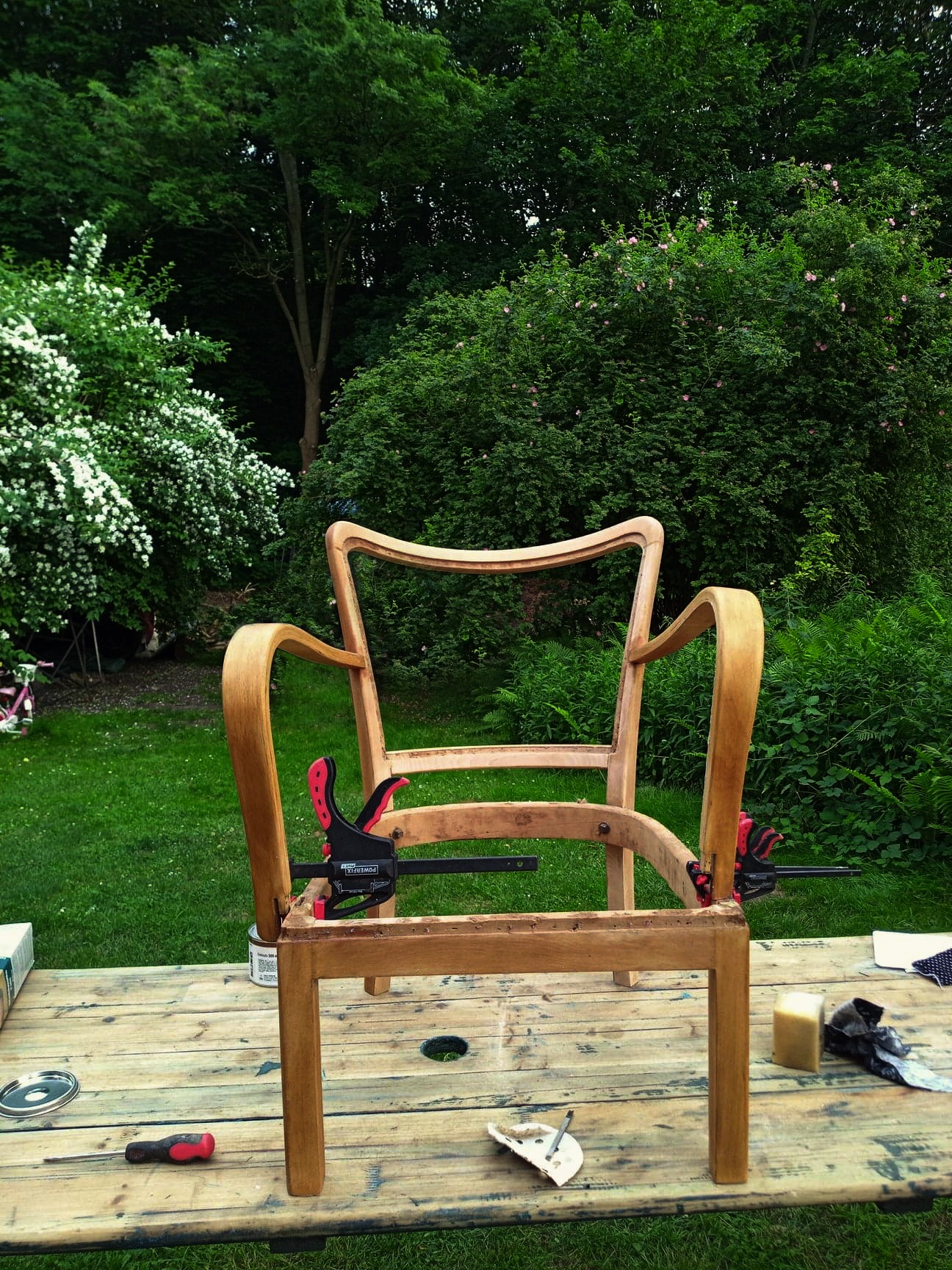 Drewniany szkielet krzesła po renowacji, zabezpieczony ściskami stolarskimi na stole warsztatowym w ogrodzie.