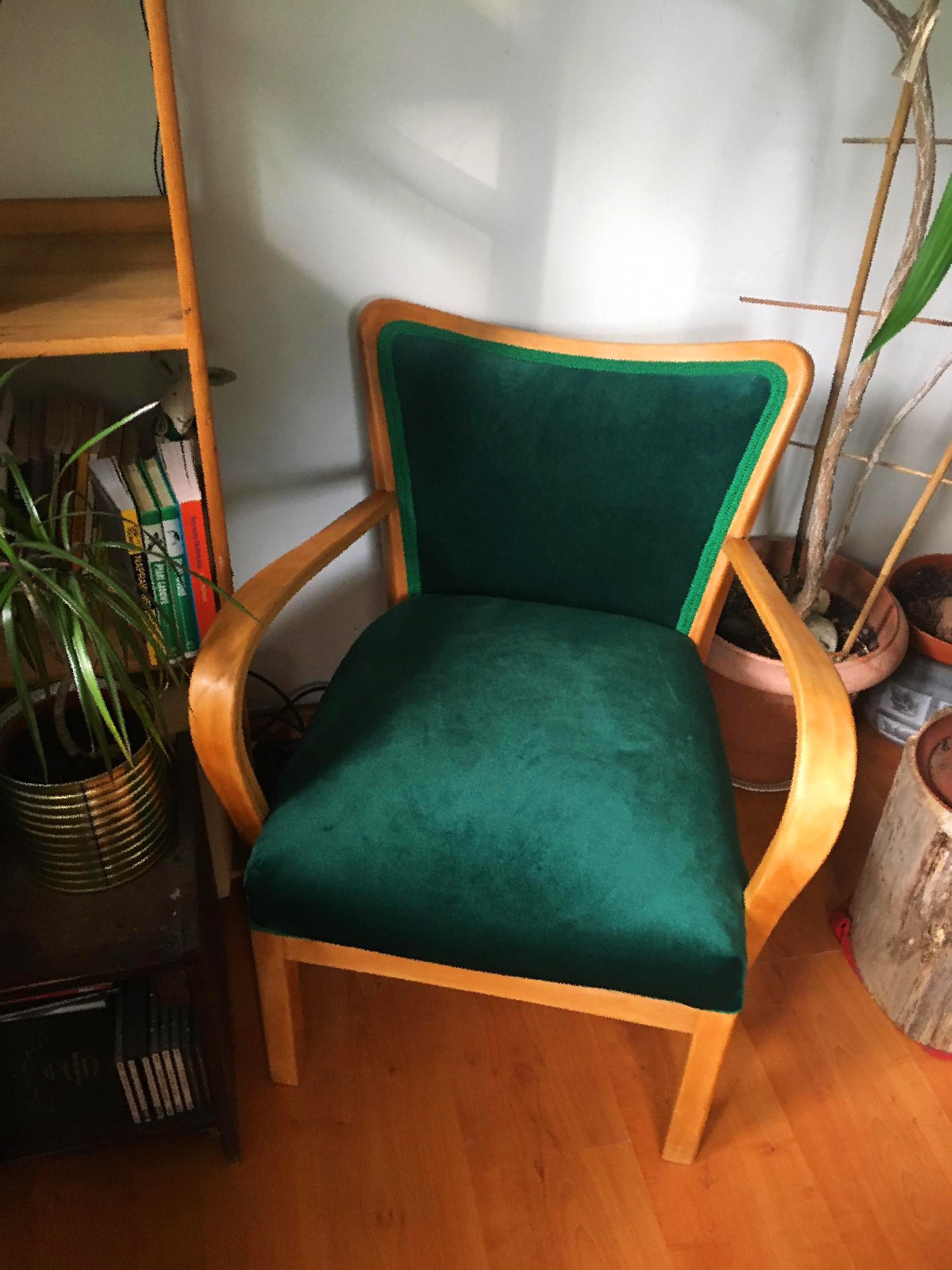 Odrestaurowany fotel w stylu retro z zieloną welurową tapicerką i drewnianą ramą, stojący na drewnianej podłodze w otoczeniu roślin i mebli.