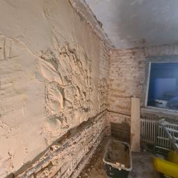 Kamil Chajecki - Ściana w trakcie prac remontowych: fragment z nałożoną grubą warstwą zaprawy, siatka wzmacniająca częściowo widoczna, odsłonięty fragment ceglanej ściany, rury instalacyjne, wiadro z resztkami...