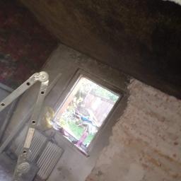 Kamil Chajecki - Poddasze w trakcie remontu, widoczna drabina oparta o ścianę z oknem, sufit częściowo pokryty ciemnym materiałem, fragment ściany z cegły oczekuje na wykończenie.