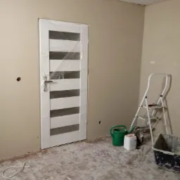 Pokój w trakcie prac wykończeniowych: ściany pomalowane na beżowo, nowa, zabezpieczona folią drzwi z poziomymi przeszkleniami, drabina malarska, wiadro i akcesoria malarskie na podłodze z resztkami...