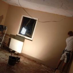 Kamil Chajecki - Pokój w trakcie remontu: przygotowanie ścian do malowania, widoczne ślady po szpachlowaniu, odsłonięta instalacja elektryczna, okno zabezpieczone folią, grzejnik, wiadro i pracownik z narzędziami...