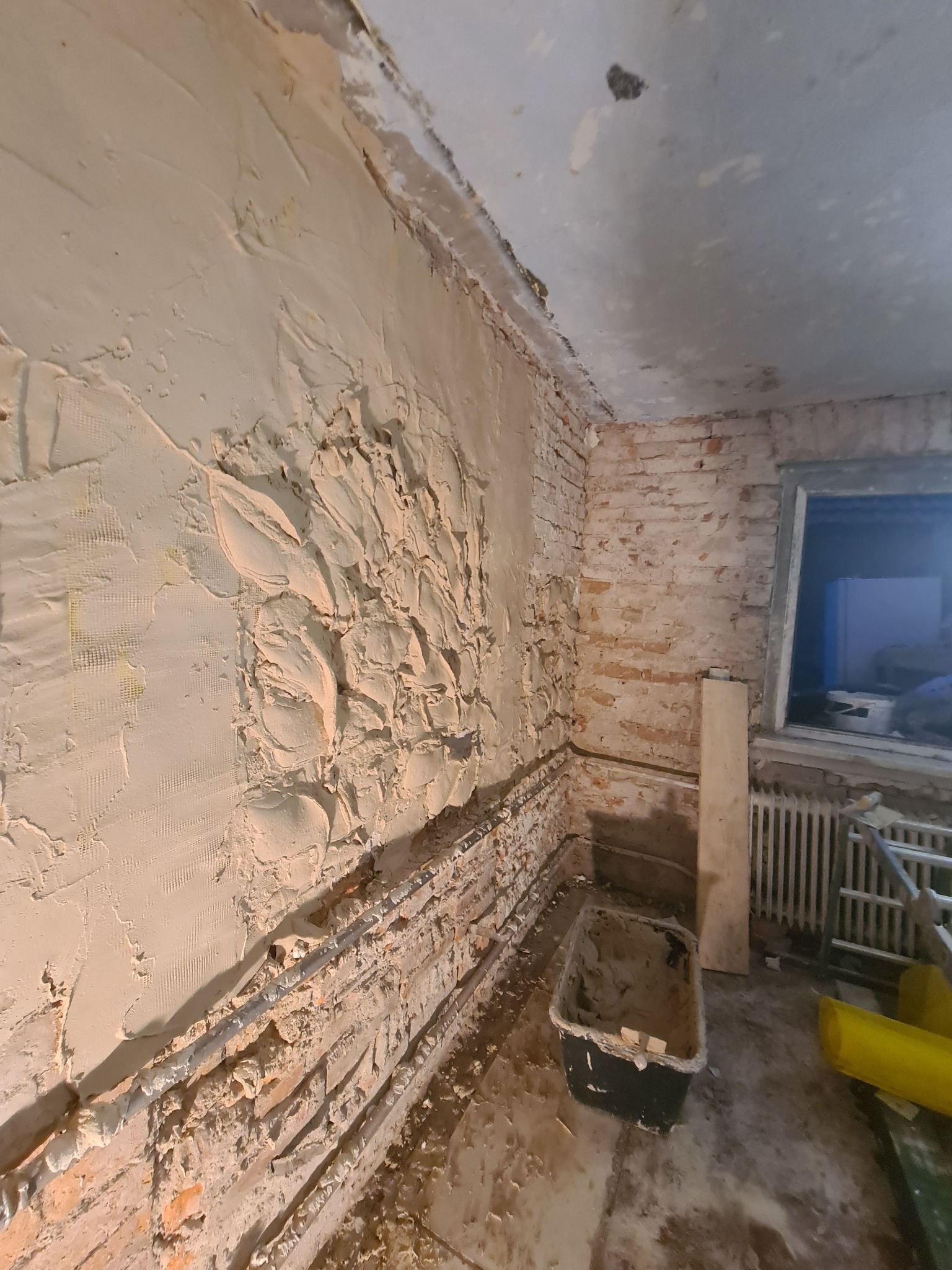 Ściana w trakcie prac remontowych: fragment z nałożoną grubą warstwą zaprawy, siatka wzmacniająca częściowo widoczna, odsłonięty fragment ceglanej ściany, rury instalacyjne, wiadro z resztkami...