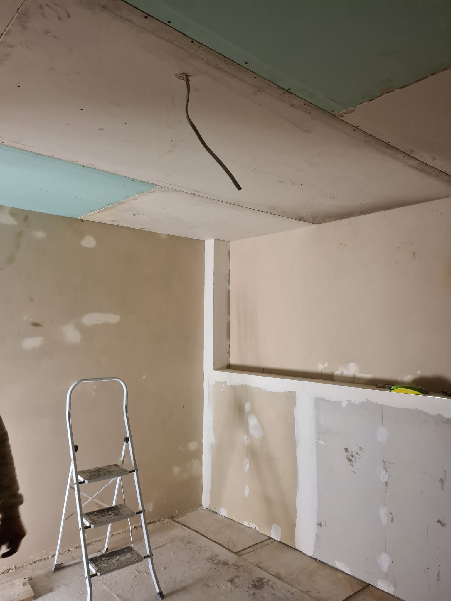 Wnętrze w trakcie montażu płyt kartonowo-gipsowych na ścianach i suficie, widoczna drabina aluminiowa, szpachlowane fragmenty ścian i wystający przewód elektryczny z sufitu.