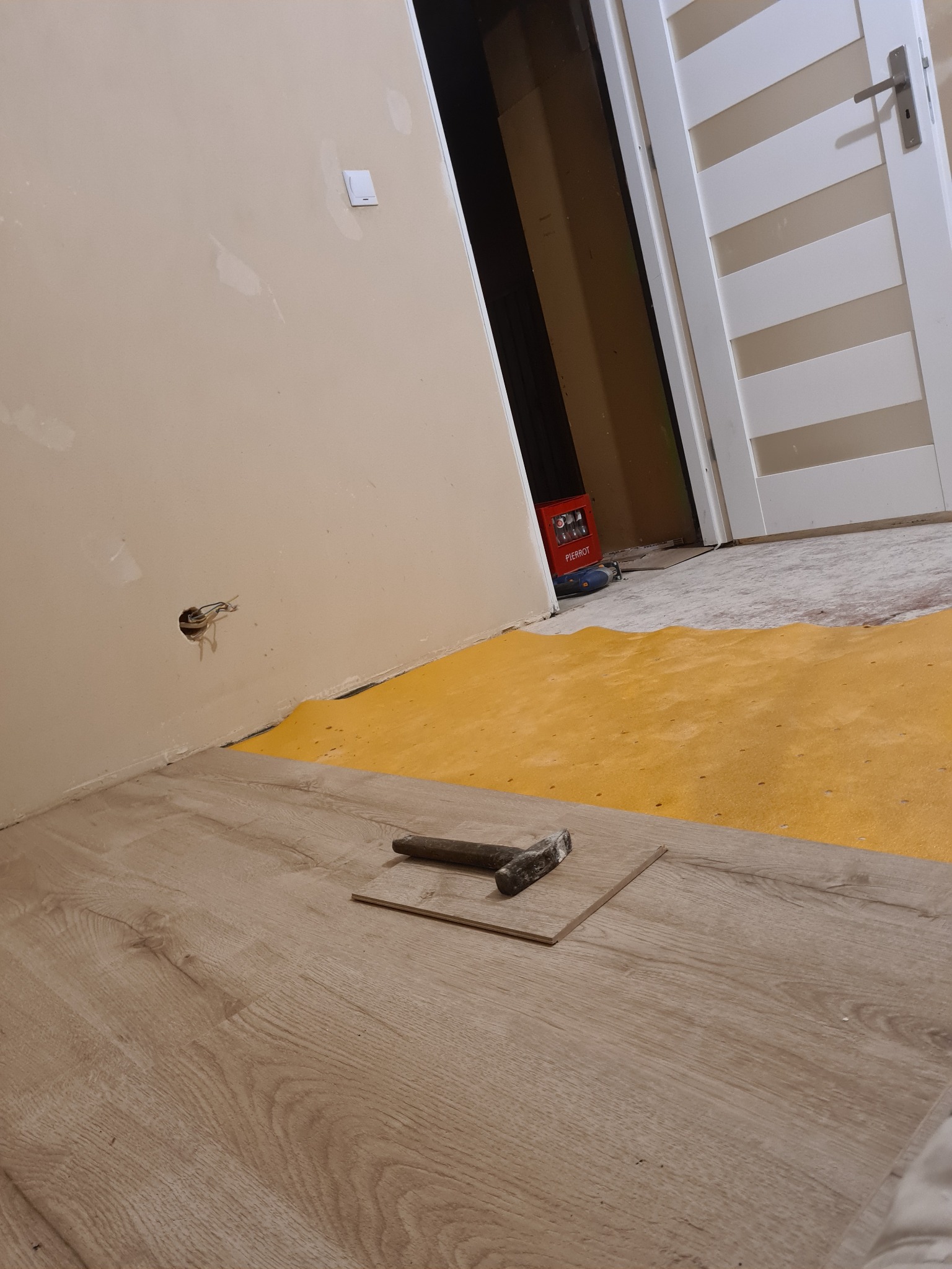 Fragment podłogi z paneli w trakcie montażu, widoczny żółty podkład, młotek i luźna deska, surowa ściana z wystającymi kablami elektrycznymi w tle.