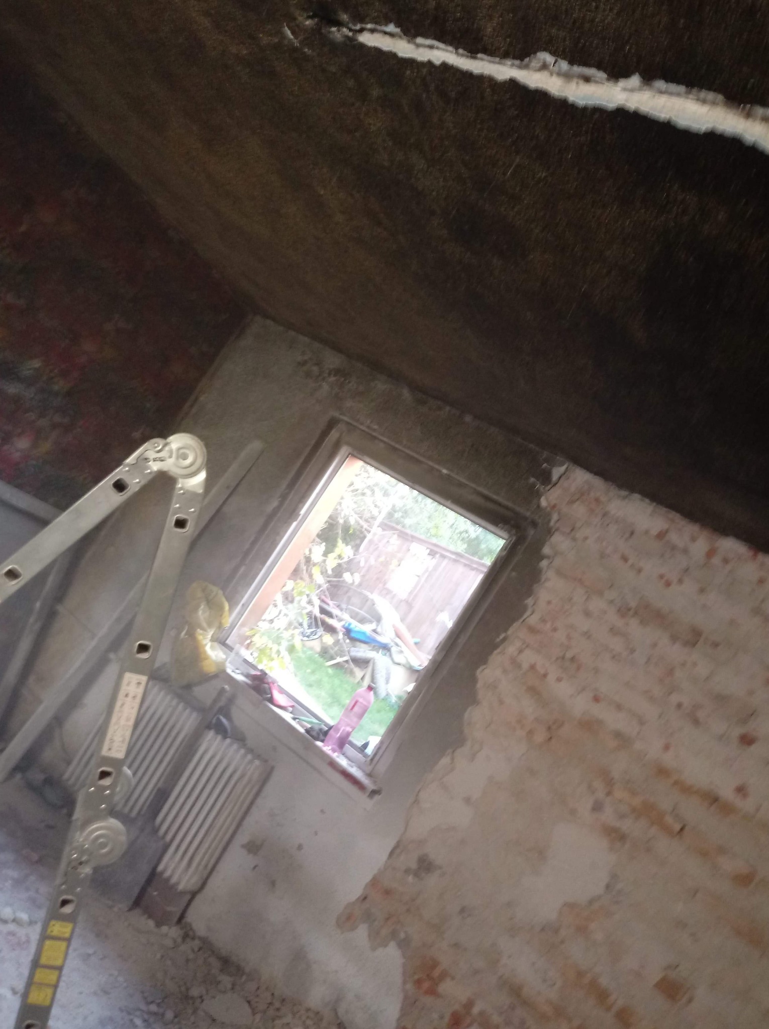 Poddasze w trakcie remontu, widoczna drabina oparta o ścianę z oknem, sufit częściowo pokryty ciemnym materiałem, fragment ściany z cegły oczekuje na wykończenie.