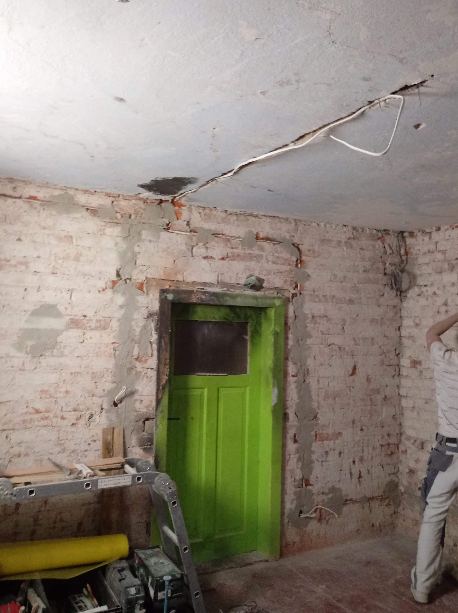 Pokój w trakcie remontu: surowa cegła z fragmentami zaprawy, zielone drzwi, widoczne kable na suficie i drabina z narzędziami.