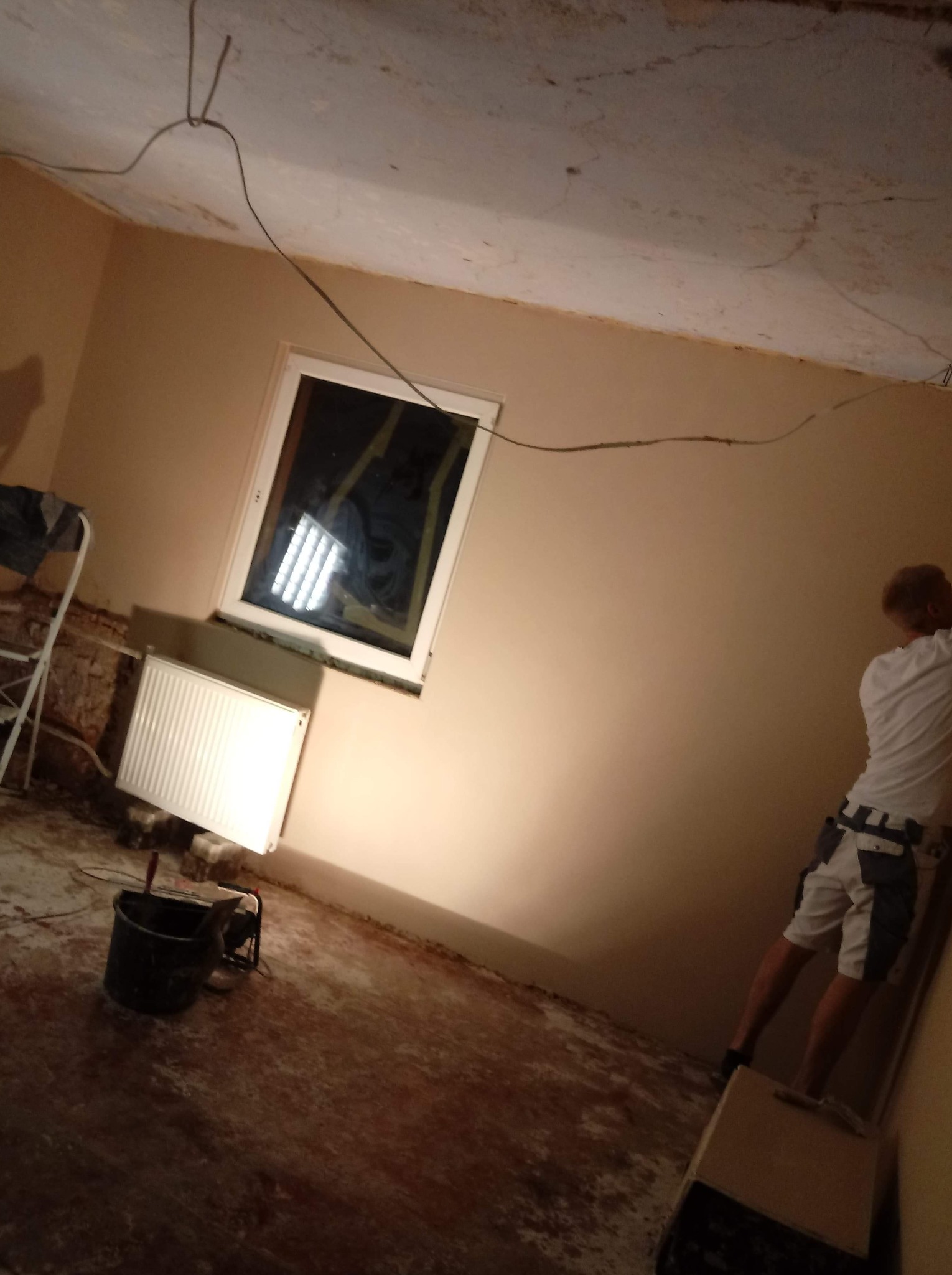 Pokój w trakcie remontu: przygotowanie ścian do malowania, widoczne ślady po szpachlowaniu, odsłonięta instalacja elektryczna, okno zabezpieczone folią, grzejnik, wiadro i pracownik z narzędziami...