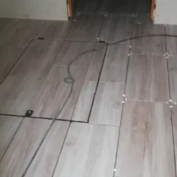 Podłoga wyłożona płytkami imitującymi drewno, z widocznymi krzyżykami dystansowymi i klapą rewizyjną z metalowym uchwytem oraz czarnym przewodem.