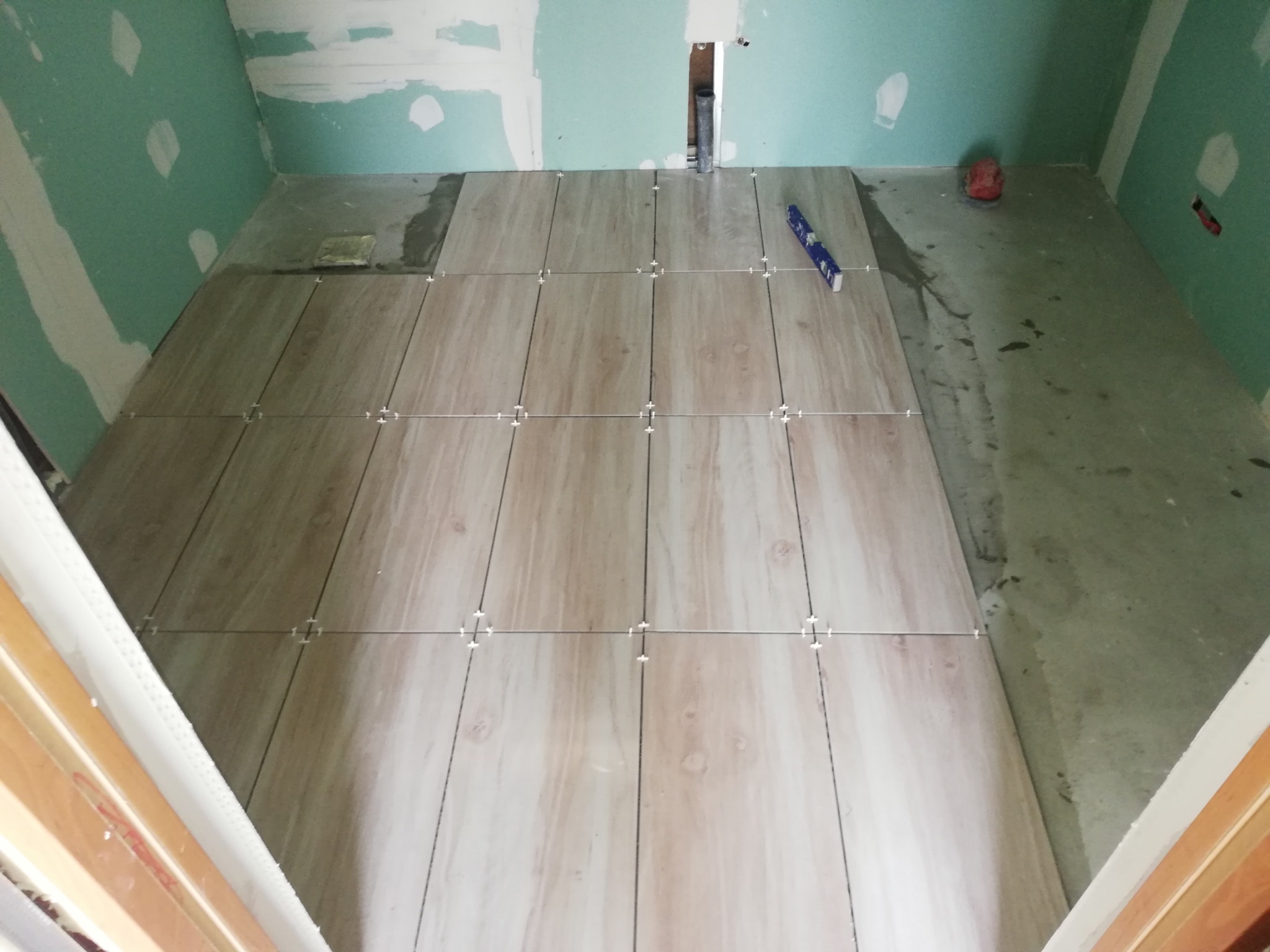 Fragment łazienki w trakcie układania podłogi z płytek imitujących drewno, widoczne krzyżyki dystansowe i poziomica na płytkach, ściany w stanie surowym.