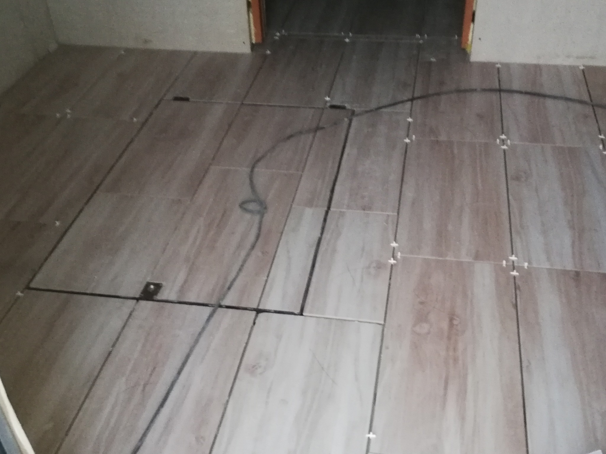 Podłoga wyłożona płytkami imitującymi drewno, z widocznymi krzyżykami dystansowymi i klapą rewizyjną z metalowym uchwytem oraz czarnym przewodem.