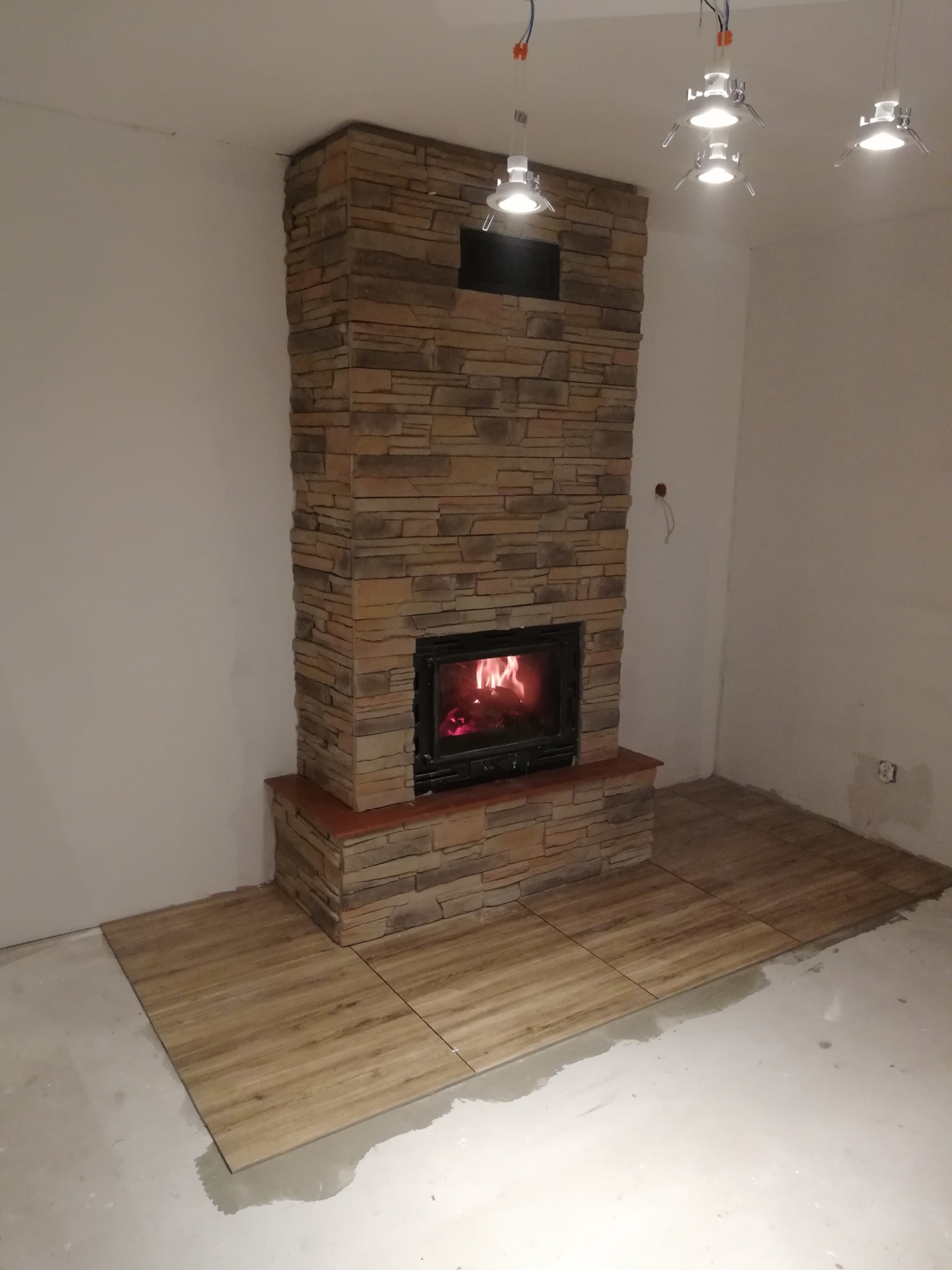 Murowany kominek z paleniskiem w trakcie wykańczania, widoczne niedokończone ułożenie paneli podłogowych i surowe ściany.
