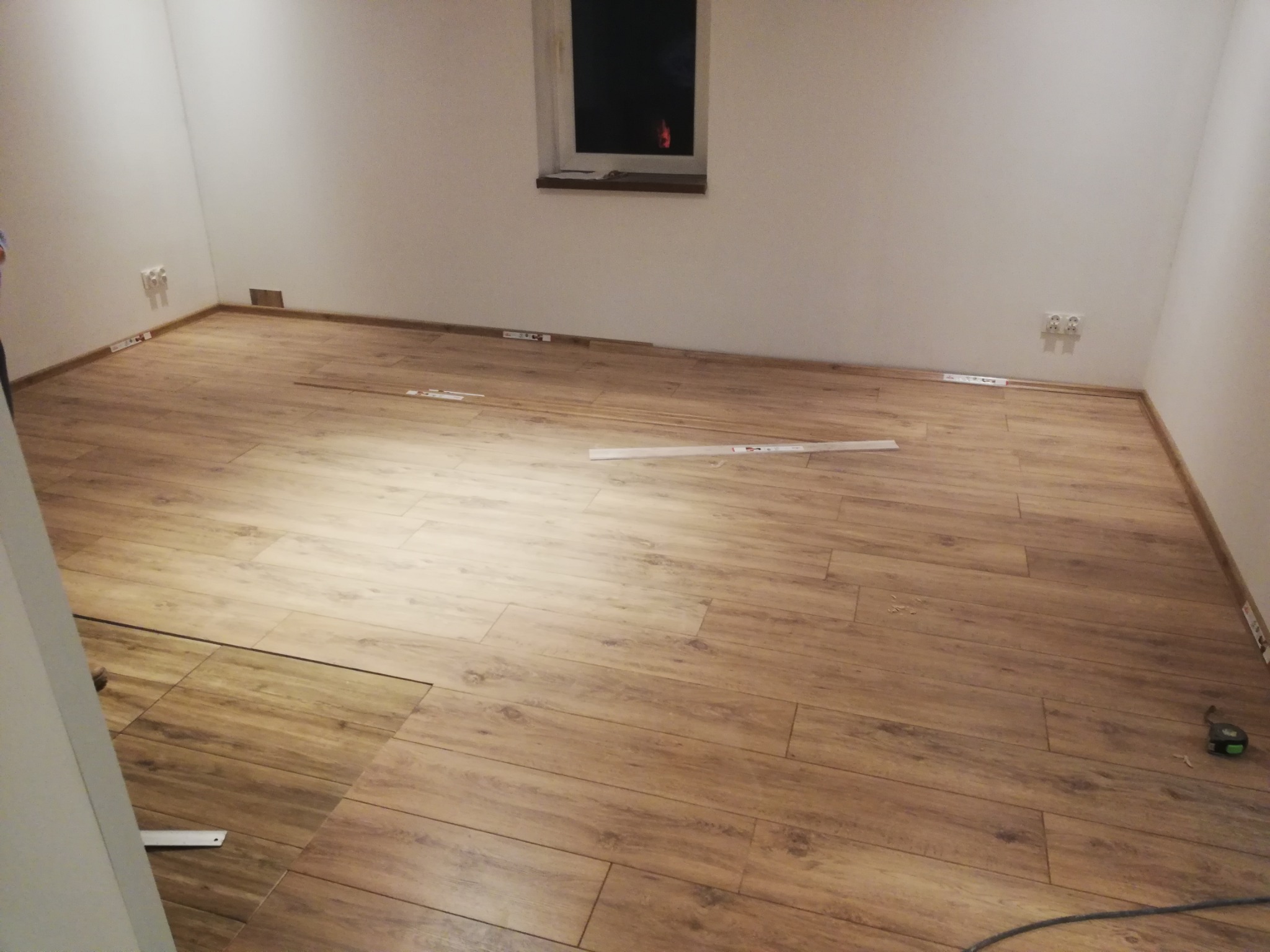 Pokój w trakcie układania paneli podłogowych o wzorze drewna, widoczne listwy przypodłogowe, poziomica i miarka.