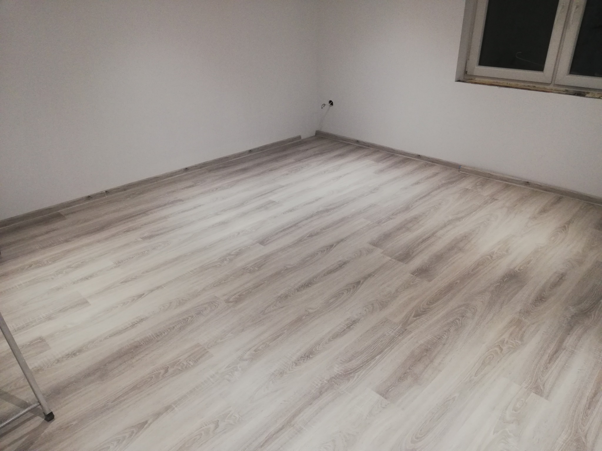 Jasne panele podłogowe imitujące drewno ułożone w pustym pokoju z białymi ścianami i oknem w tle, widoczne gniazdka elektryczne.