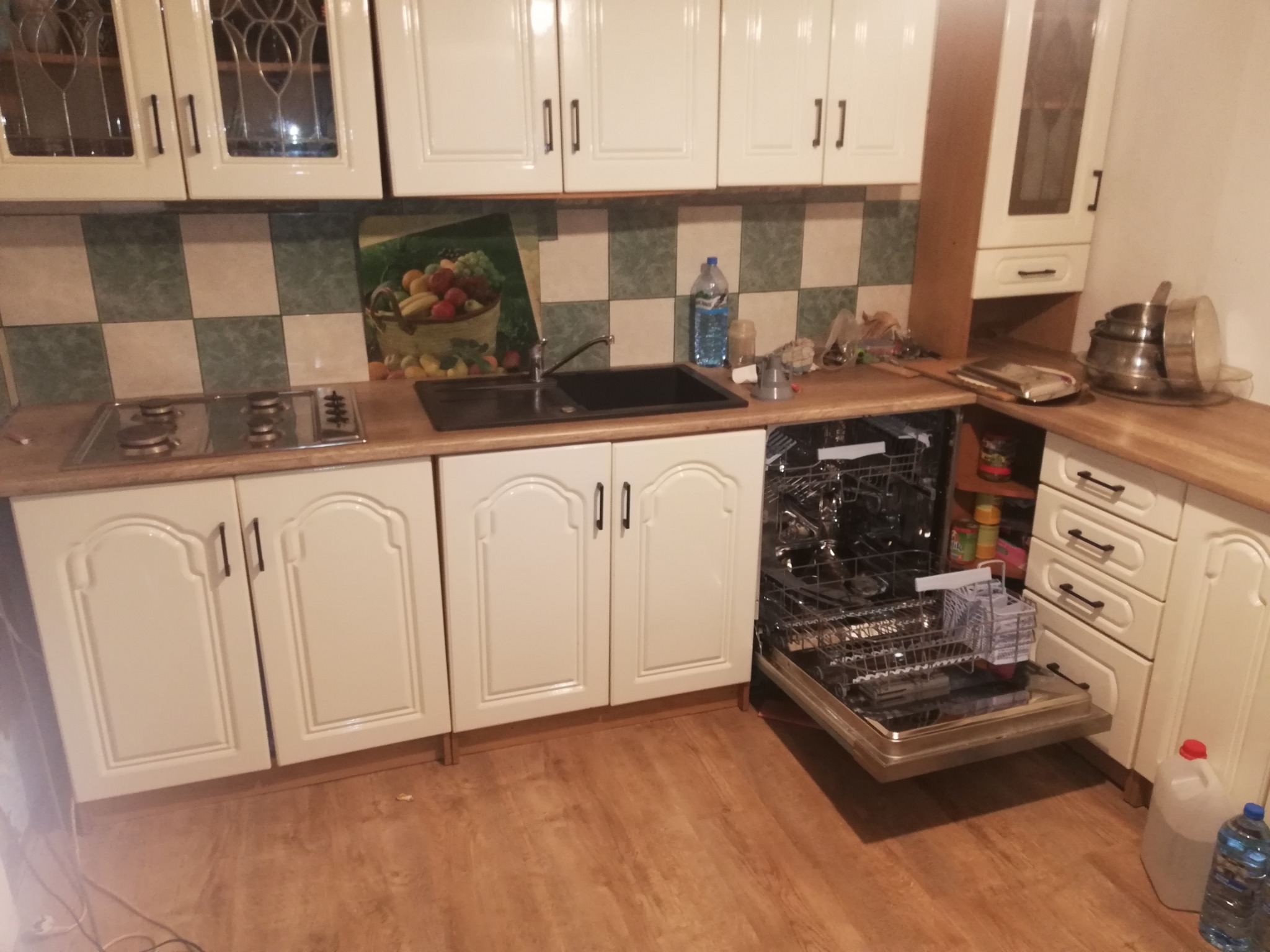 Kuchnia z kremowymi szafkami, kuchenką gazową i otwartą zmywarką, na blacie butelka wody i akcesoria kuchenne.