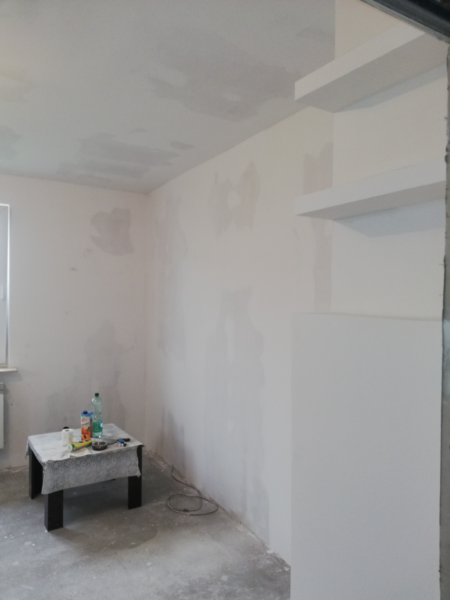 Pokój w trakcie przygotowania do malowania: ściany po szpachlowaniu, widoczne fragmenty płyty karton gips, mały stolik z materiałami i napojami w rogu pomieszczenia, surowy beton na podłodze.