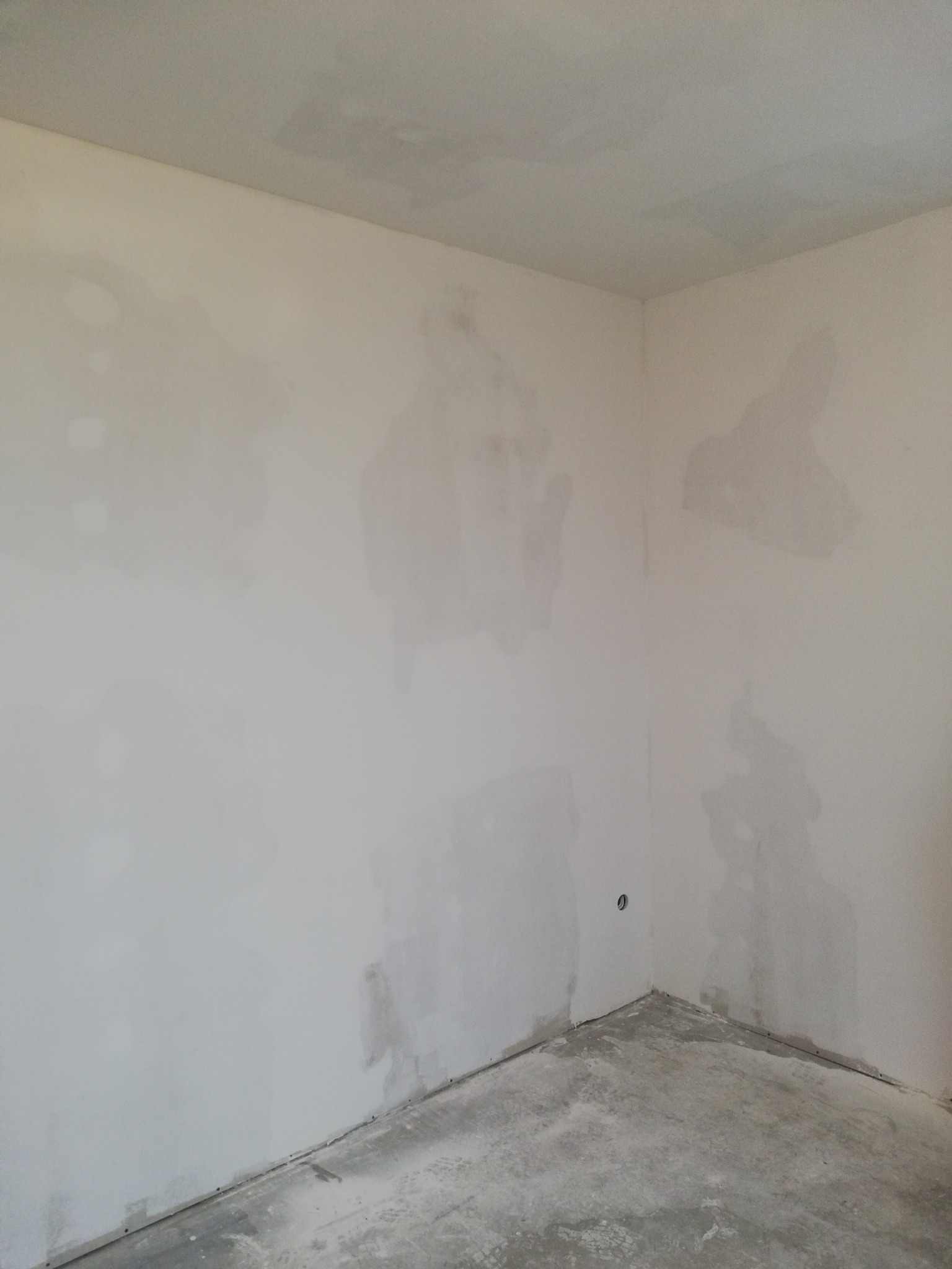 Pokój w trakcie prac przygotowawczych do malowania, ściany z widocznymi plamami po szpachlowaniu i gruntowaniu, surowa betonowa podłoga, gniazdko elektryczne.