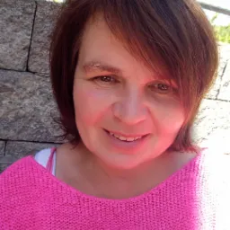 Portret uśmiechniętej kobiety w różowym swetrze na tle kamiennej ściany i zieleni ogrodu, z widoczną latarnią.