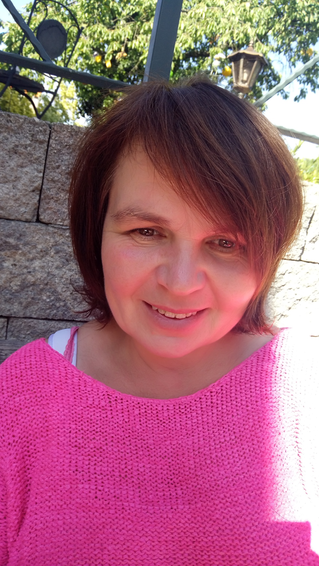 Portret uśmiechniętej kobiety w różowym swetrze na tle kamiennej ściany i zieleni ogrodu, z widoczną latarnią.