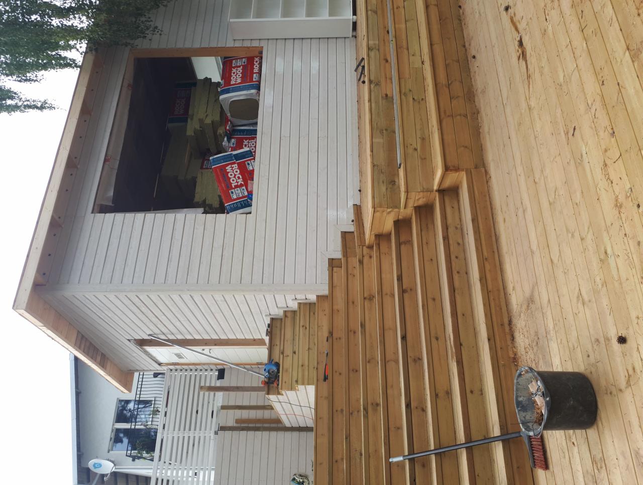 Wznoszenie drewnianego tarasu przy białym domu z oknem, w którym widoczne są paczki wełny mineralnej; widok z dołu, kubeł i szczotka na podłodze.