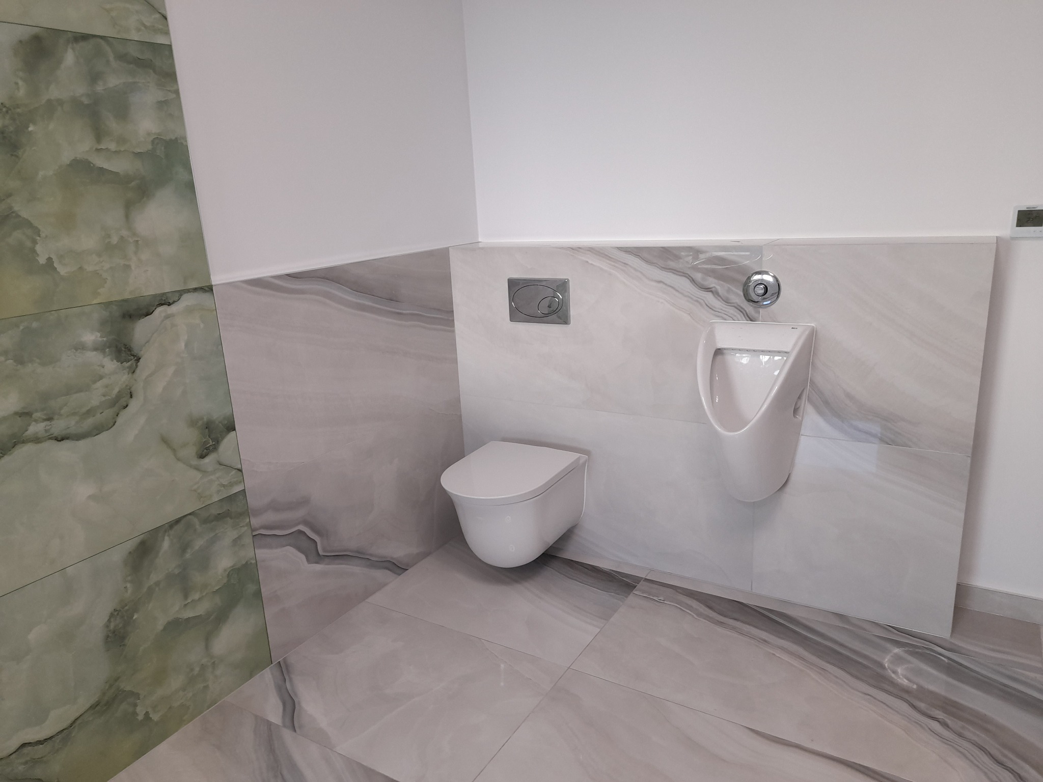 Nowoczesna łazienka z podwieszaną toaletą i pisuaru, wykończona płytkami imitującymi marmur w odcieniach szarości i zieleni, z widocznym przyciskiem spłukiwania i termostatem.