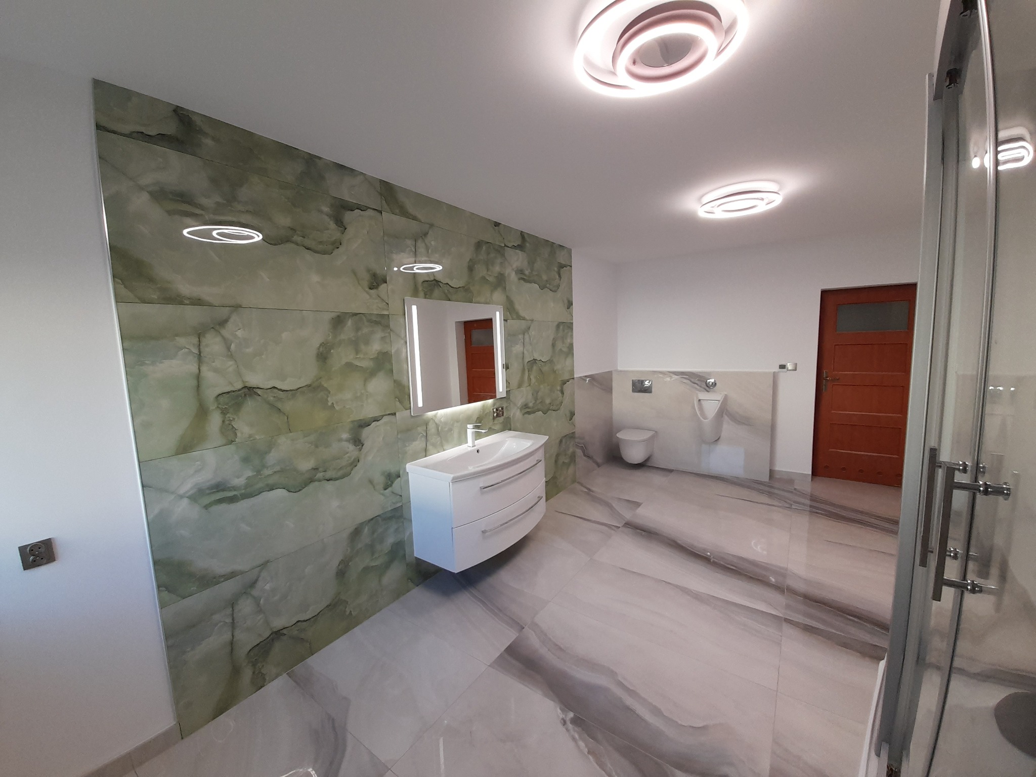 Elegancka łazienka z zielonymi płytkami imitującymi marmur na ścianie, wisząca biała szafka z umywalką, podświetlane lustro i dekoracyjne, okrągłe lampy sufitowe. Widoczne również toaleta, pisuar...