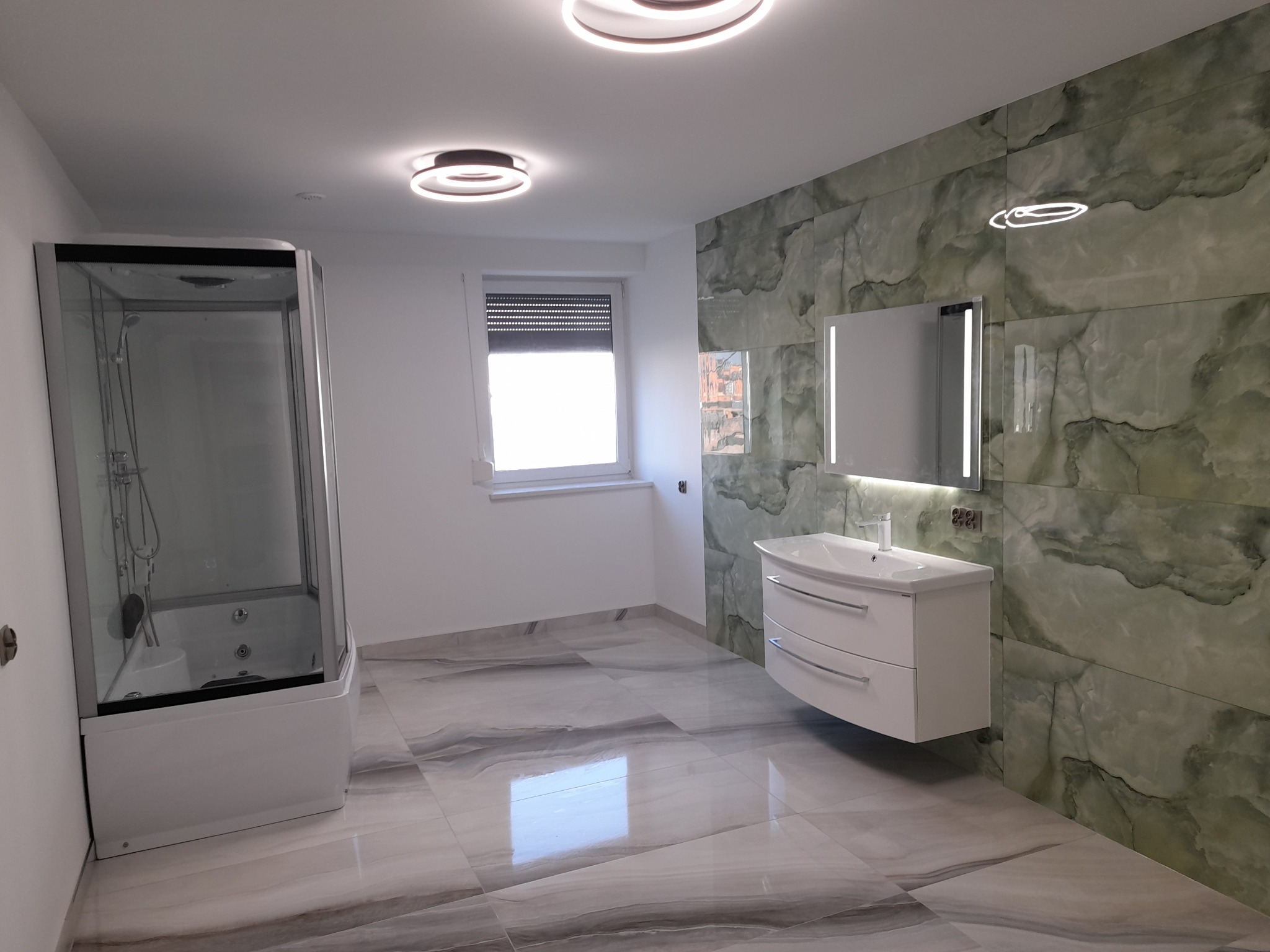 Wykończona łazienka z kabiną prysznicową z hydromasażem, umywalką z szafką wiszącą i zielonymi płytkami imitującymi marmur na ścianie.