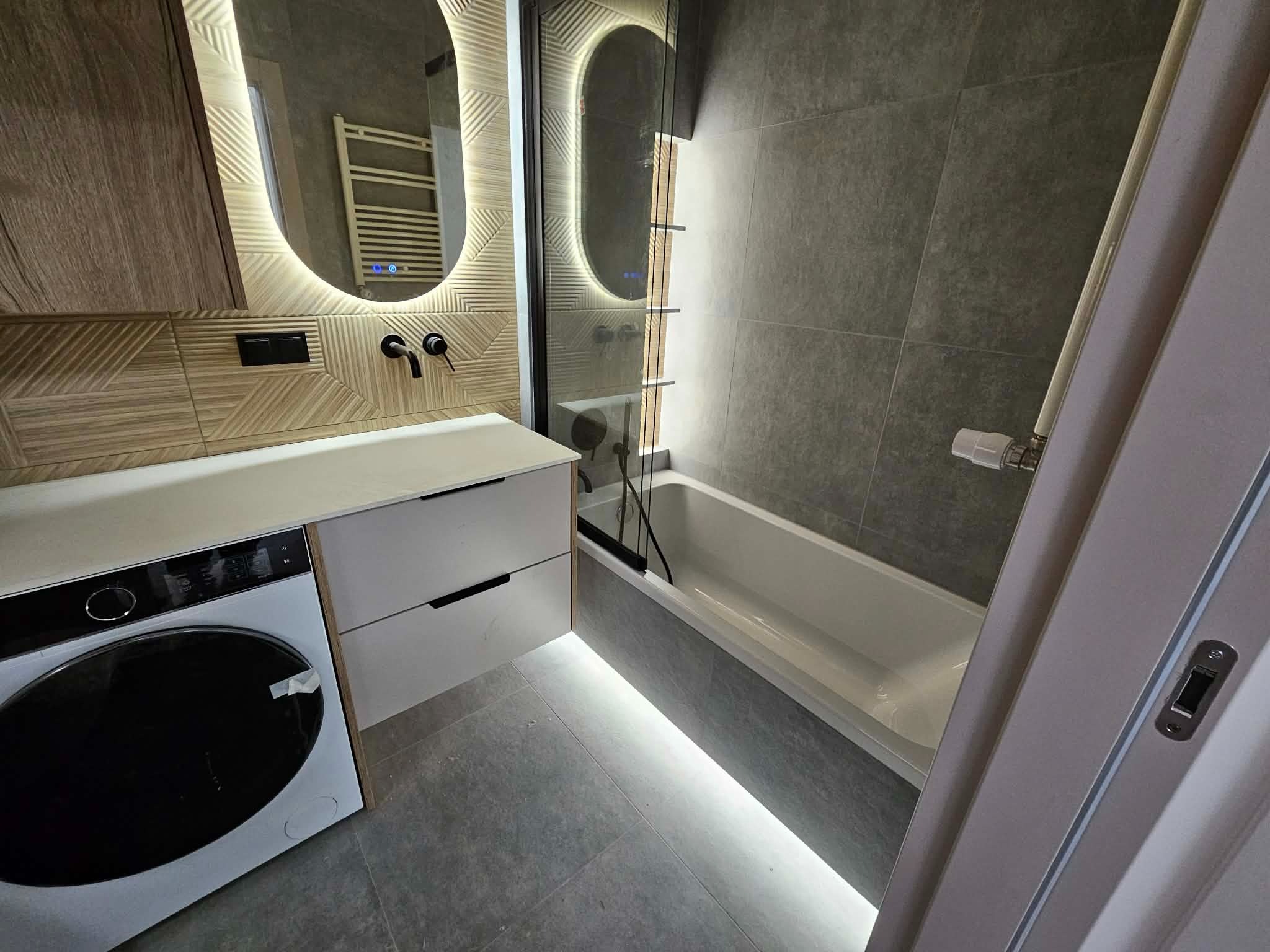 Nowoczesna łazienka z podświetlaną wanną, umywalką z szafką i pralką. Szare płytki, lustro z oświetleniem LED i armatura w czarnym macie tworzą elegancki wystrój.