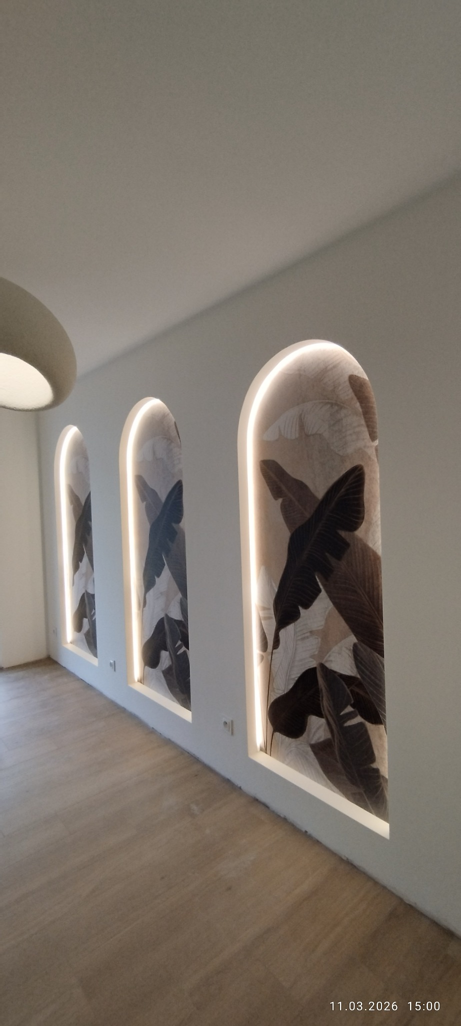 Trzy wnęki w kształcie łuków z podświetleniem LED i tapetą z motywem liści bananowca na białej ścianie. Parkiet na podłodze. Nowoczesny design.