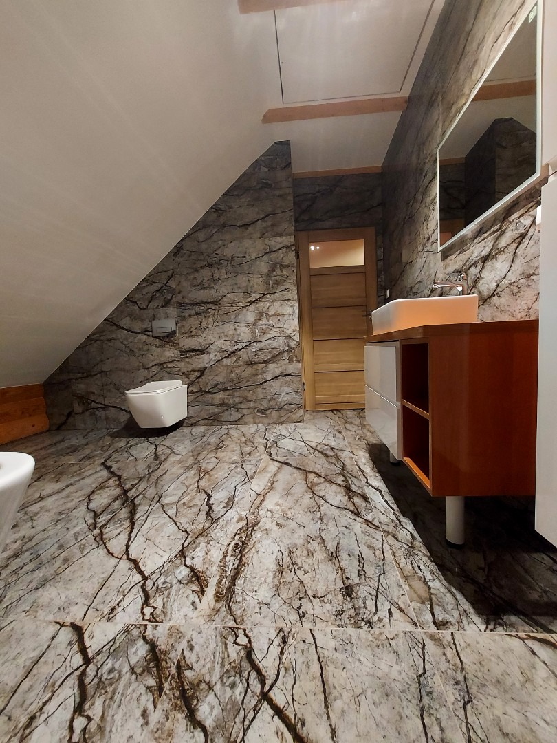 Wnętrze łazienki na poddaszu z marmurkowymi płytkami na podłodze i ścianach, wisząca toaleta, umywalka z szafką i drewniane drzwi.