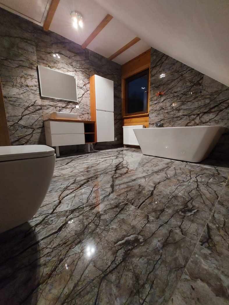 Nowoczesna łazienka na poddaszu z marmurowymi płytkami na ścianach i podłodze, biała wolnostojąca wanna, toaleta i szafka z umywalką.