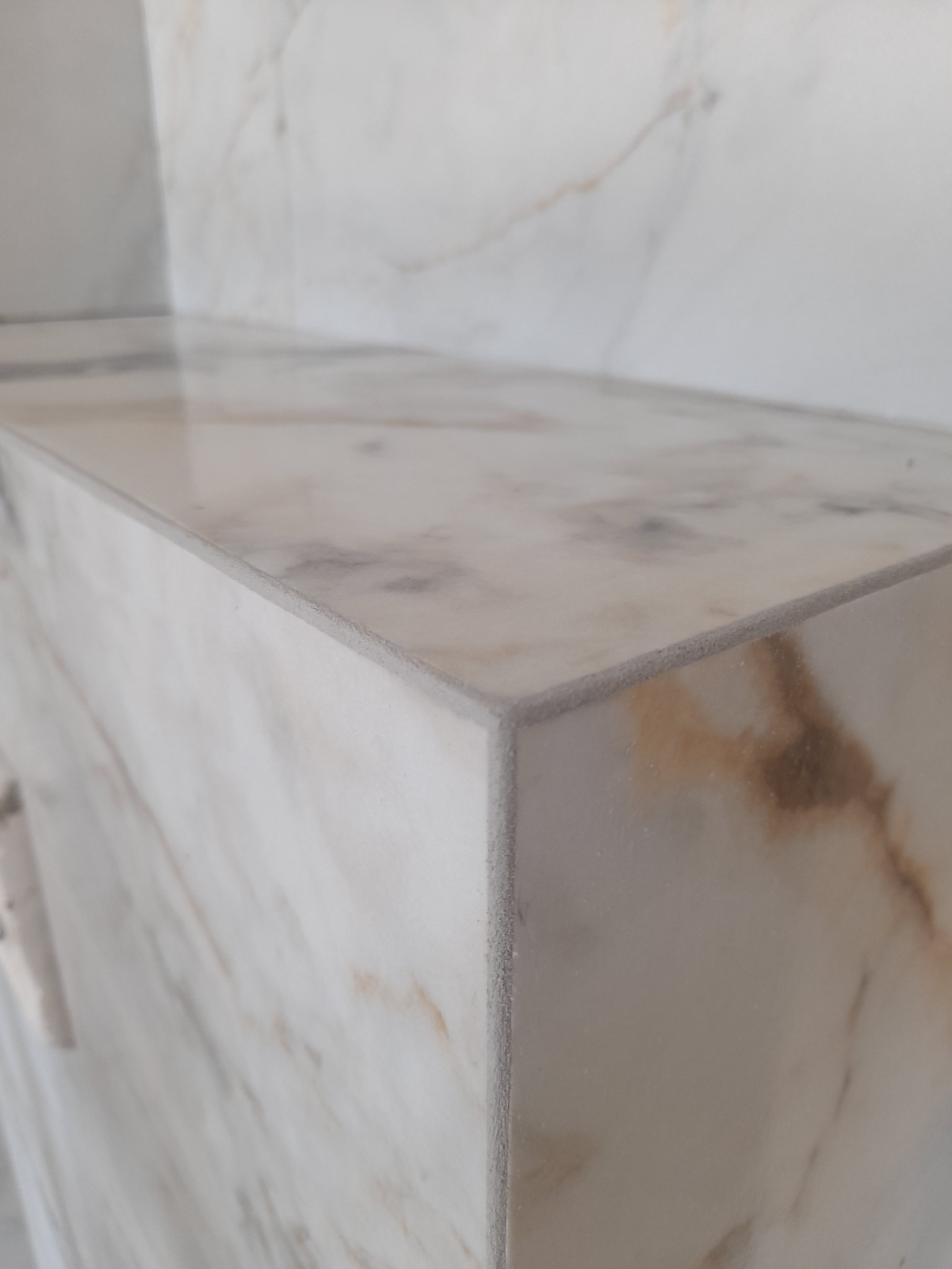 Wykończony narożnik półki z płytek imitujących marmur, z widoczną szarą krawędzią cięcia i fugą, na jasnym tle.