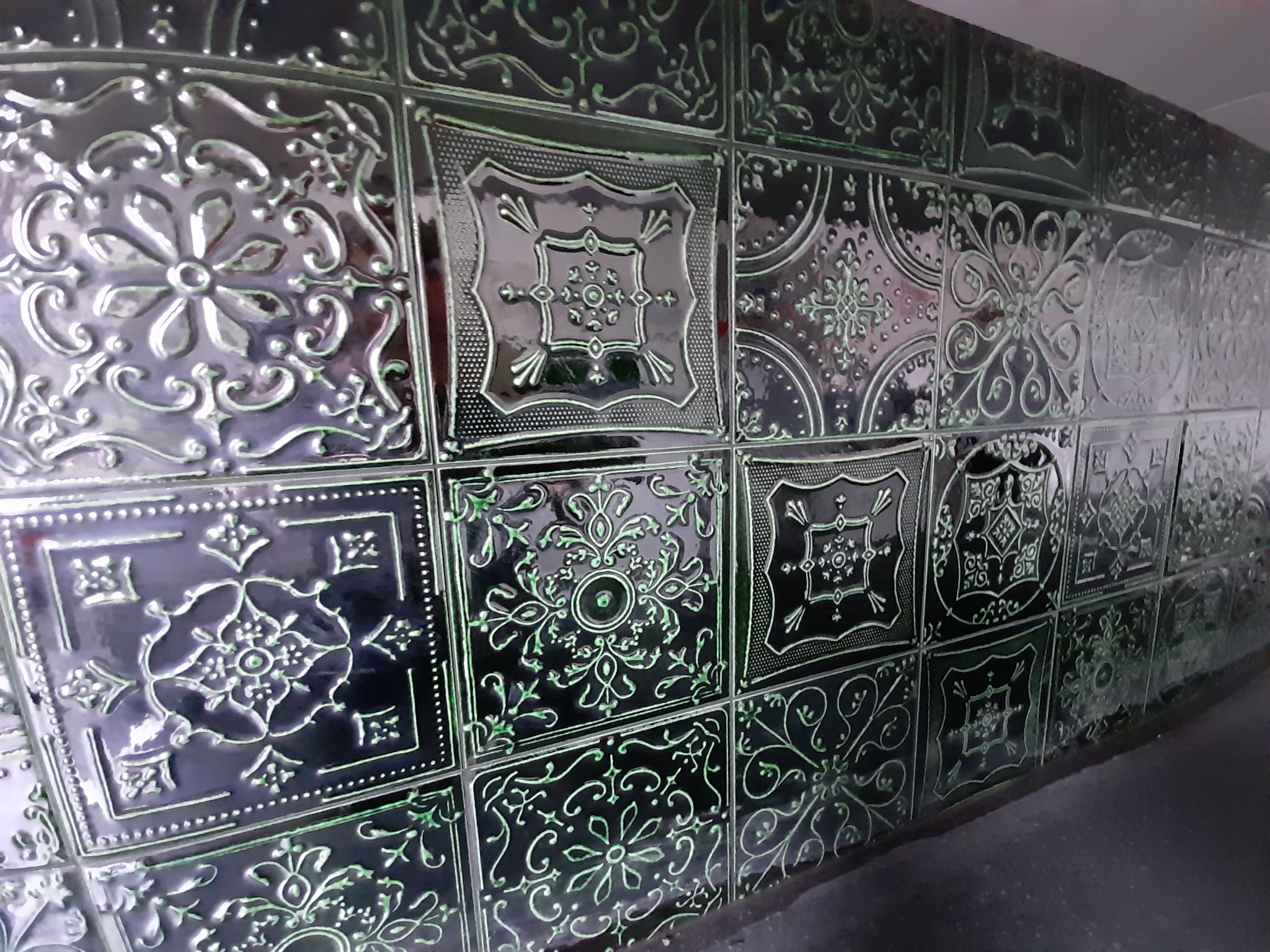 Ściana wyłożona ciemnymi, błyszczącymi płytkami ceramicznymi z wytłaczanymi, symetrycznymi wzorami kwiatowymi i geometrycznymi, ułożonymi pod kątem.