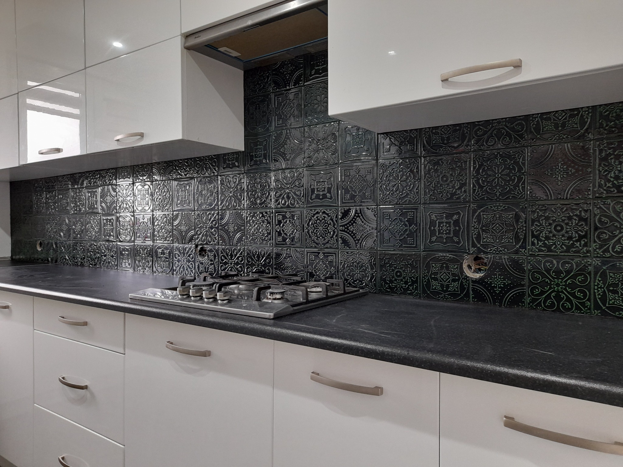 Nowoczesna kuchnia z białymi szafkami i czarnym blatem, ozdobna czarna glazura z orientalnymi wzorami na ścianie nad kuchenką gazową.