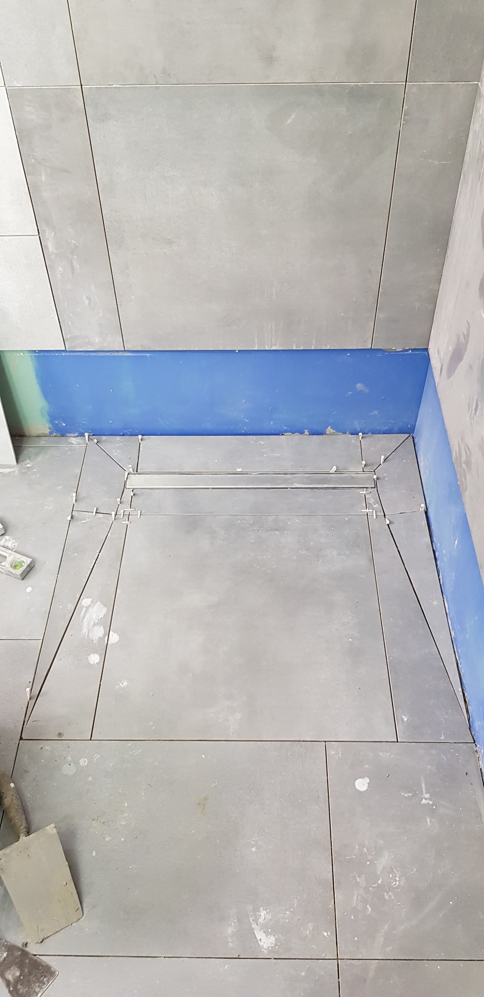 Ułożone szare płytki podłogowe w łazience z liniowym odpływem, widoczne krzyżyki dystansowe i ślady zaprawy, ściany zabezpieczone niebieską folią w płynie.